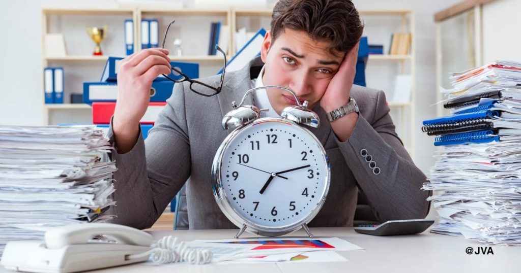 Seorang pemilik bisnis laki-laki terlihat lelah dan stres (burnout) di meja kerja yang penuh tumpukan dokumen operasional dan jam weker besar, menggambarkan pentingnya delegasi ke virtual assistant.