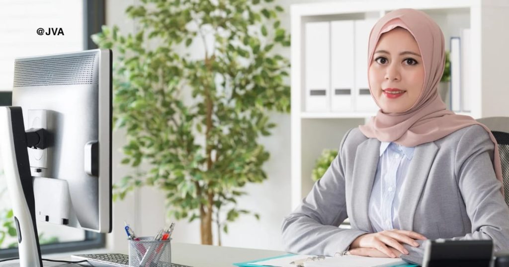 Seorang wanita berhijab dengan seragam bisnis modern sedang duduk di meja kerja kantor, mewakili profesionalisme seorang Virtual Assistant yang siap menangani tugas operasional.