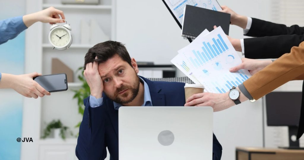 Seorang pria yang kewalahan dan frustrasi memegang kepalanya di meja kantornya, dikelilingi oleh banyak tangan yang memberinya tugas berbeda.
