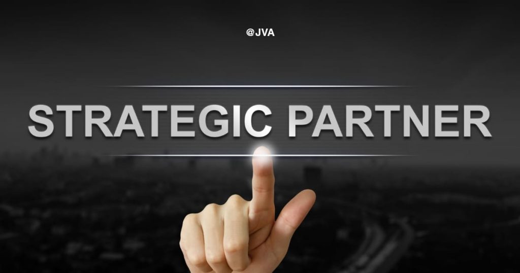 Tangan seseorang yang sedang menunjuk ke arah teks 'STRATEGIC PARTNER' yang bercahaya di latar belakang perkotaan yang gelap dan dramatis.