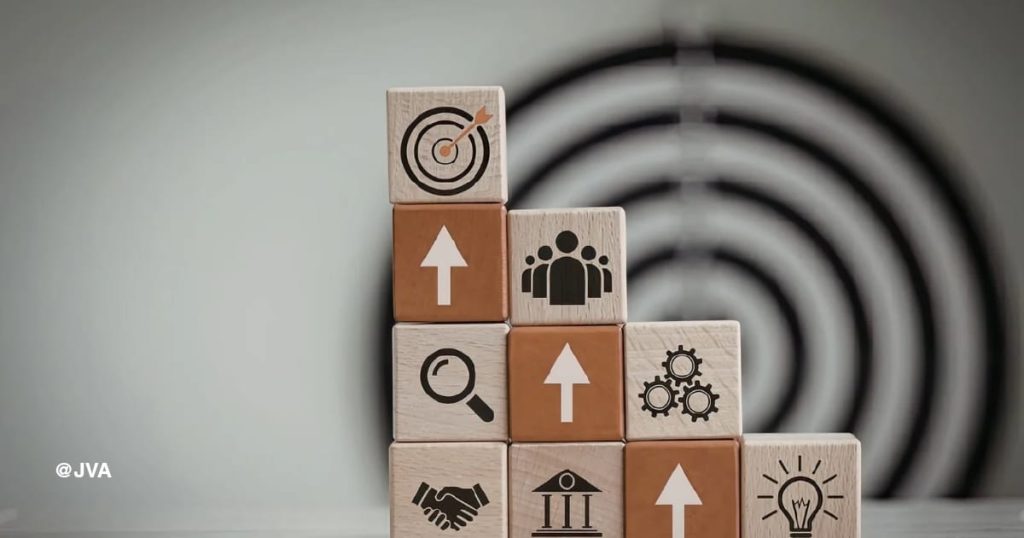 Susunan balok kayu membentuk tangga menuju target dengan ikon bisnis seperti pertumbuhan, pencarian, kemitraan, dan ide, melambangkan sistem JasaVirtualAssistantID.