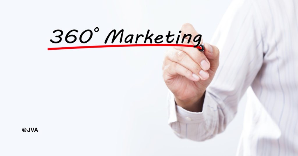 Tangan seorang profesional sedang menulis istilah '360° Marketing' dengan spidol hitam di latar belakang putih bersih.