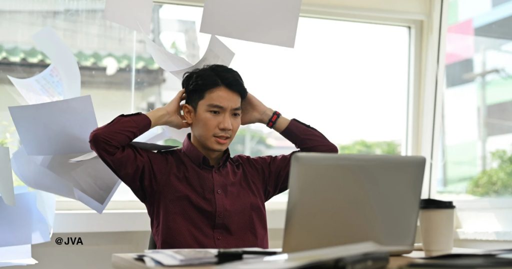 Seorang pemilik bisnis pria tampak stres di depan laptop dengan kertas-kertas berantakan, menggambarkan kesalahan merekrut virtual assistant yang tidak efektif.